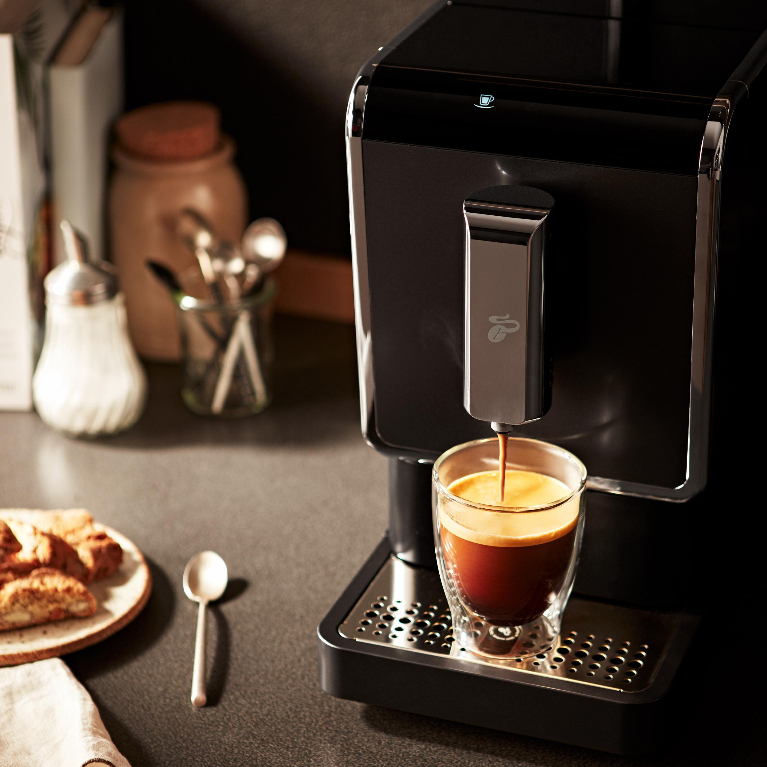 Eine schwarze Kaffeemaschine, die Kaffee in ein Glas ausgibt. Ein Teller mit Biscotti und ein Löffel sind in der Nähe.