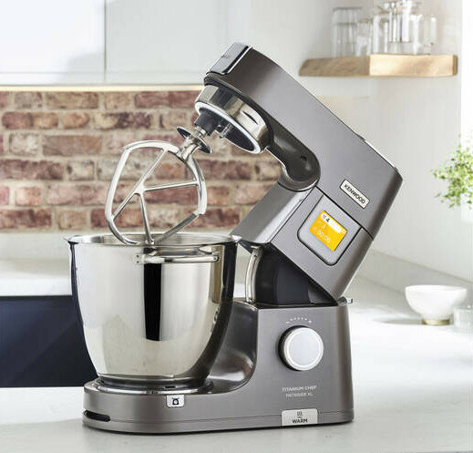 Szary robot kuchenny Kenwood titanium chef na białym blacie w pobliżu ceglanej ściany.