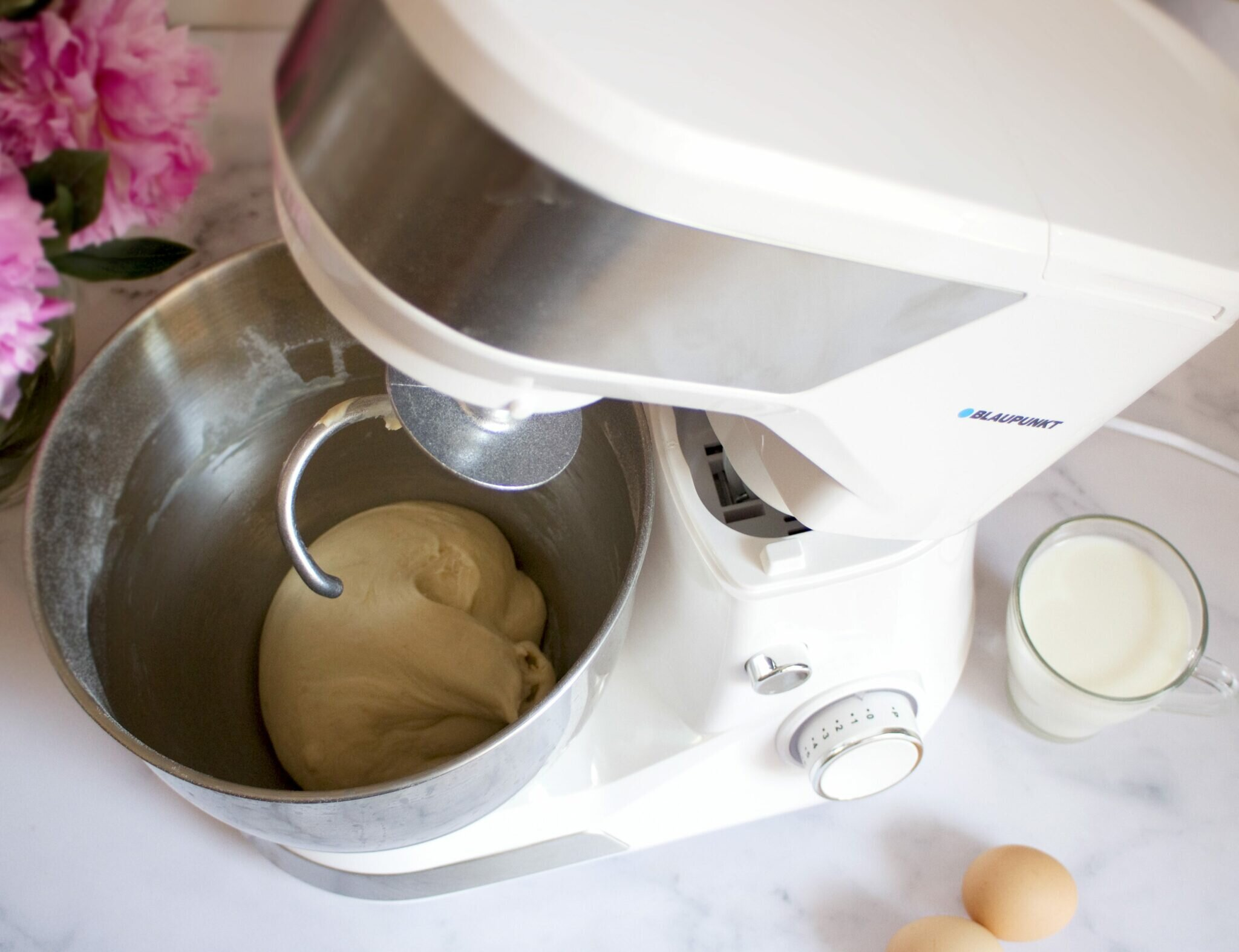 Biały i srebrny mikser stojący z ciastem i składnikami. Kwiaty w tle.