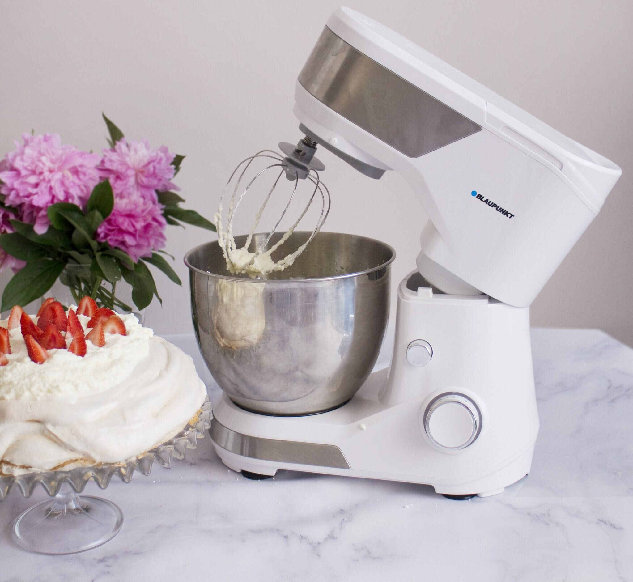 Biały mikser stojący z misą, trzepaczką i ciastem z truskawkami, obok kwiatów.