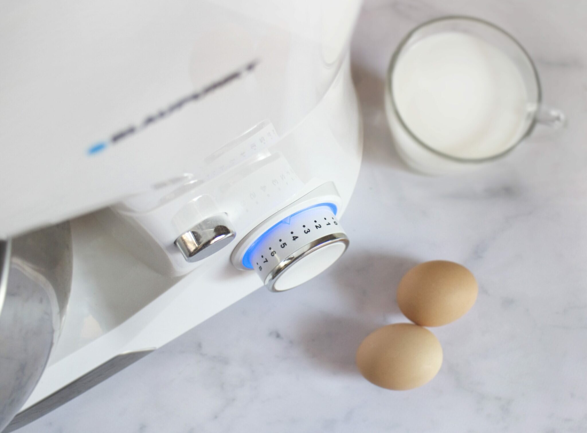 Biały mikser stojący z pokrętłem, dwoma jajkami i szklanką mleka na marmurowej powierzchni.