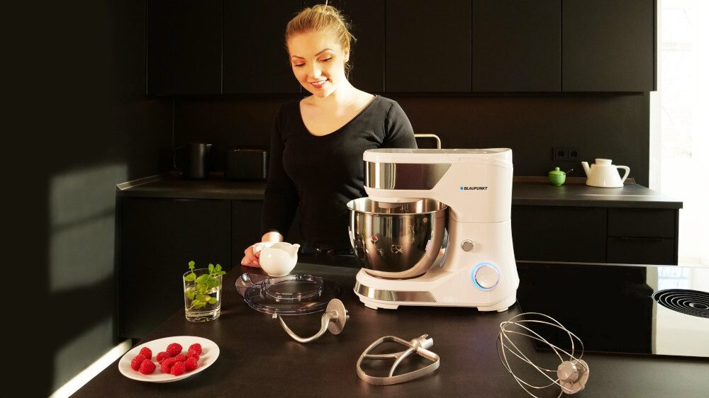 Kobieta w kuchni z mikserem. Składniki na stole. Czarne szafki w tle.