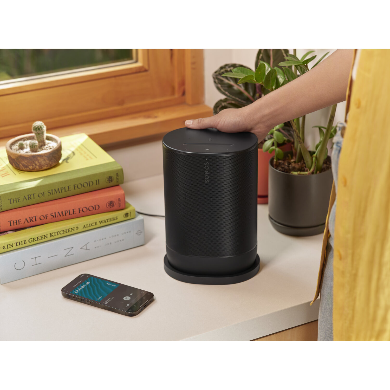 Black speaker, books, and phone on a white surface. A hand is touching the speaker.