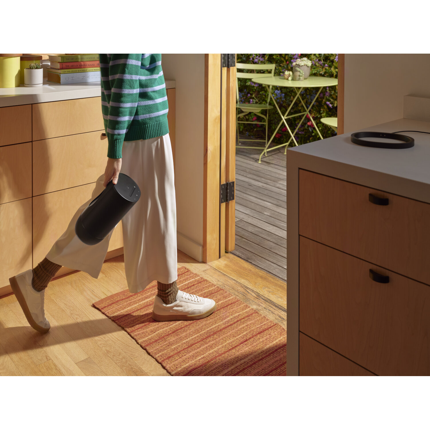 A person in a green sweater and white pants walking through a doorway, holding a black cylinder.