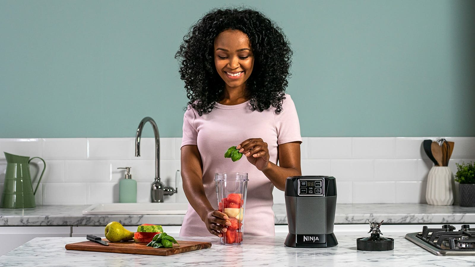 Uśmiechnięta kobieta dodająca bazylię do blendera z arbuzem i innymi składnikami na stole.