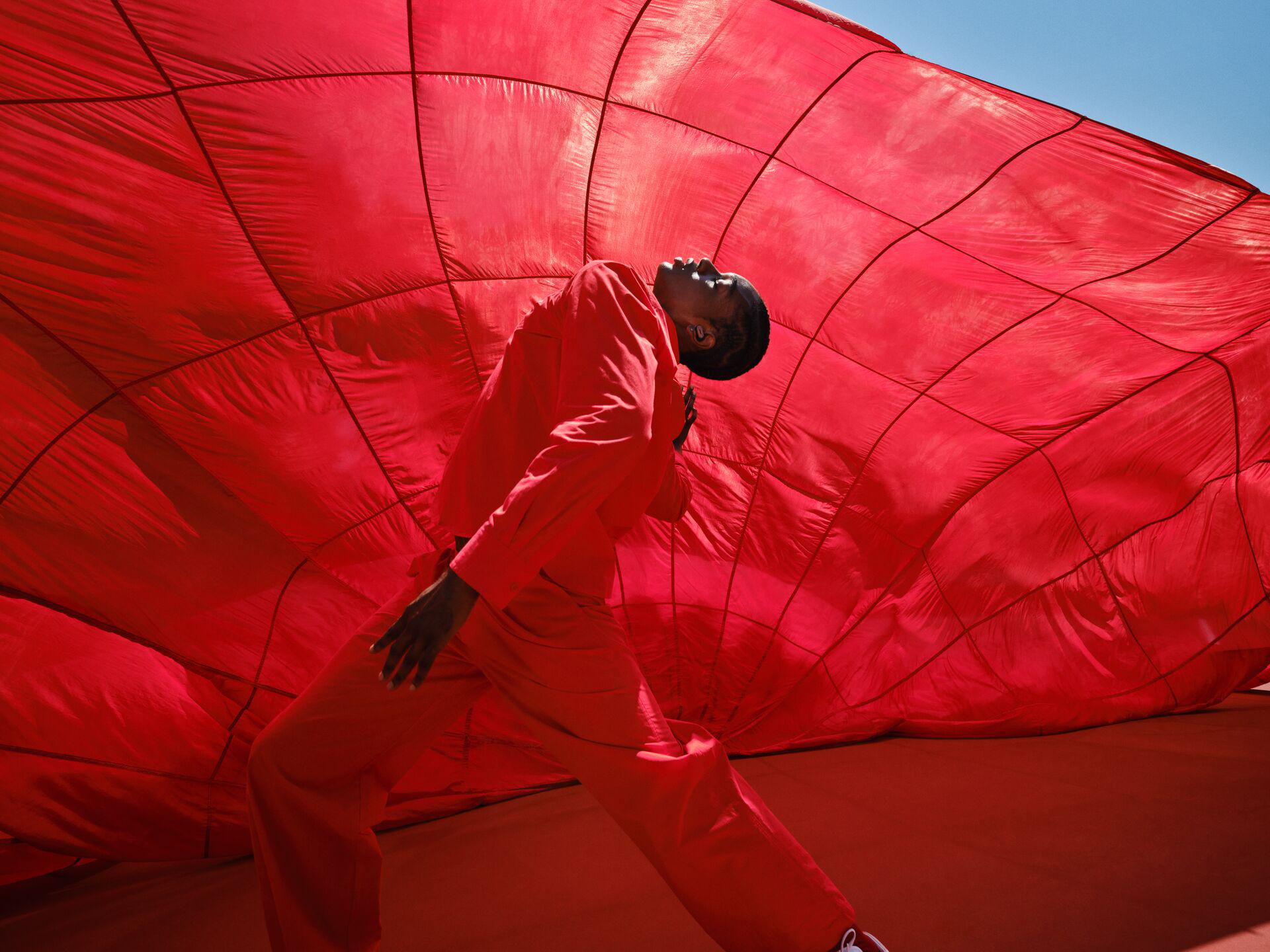 Eine Person in rotem Anzug lehnt sich an einen großen roten Ballon. Der Hintergrund ist blauer Himmel und roter Ballon.