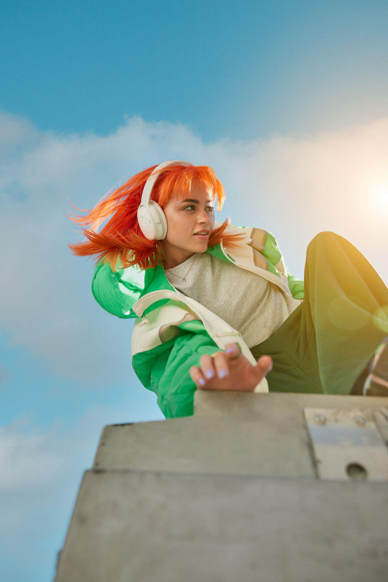 Eine Frau mit orangefarbenen Haaren und weißen Kopfhörern blickt nach rechts, vor blauem Himmel.