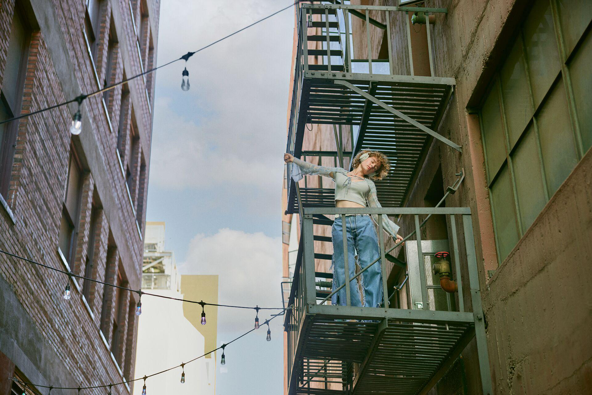 Frau auf Feuerleiter, Kopfhörer, in Gasse. Gebäude und Lichterketten im Hintergrund.