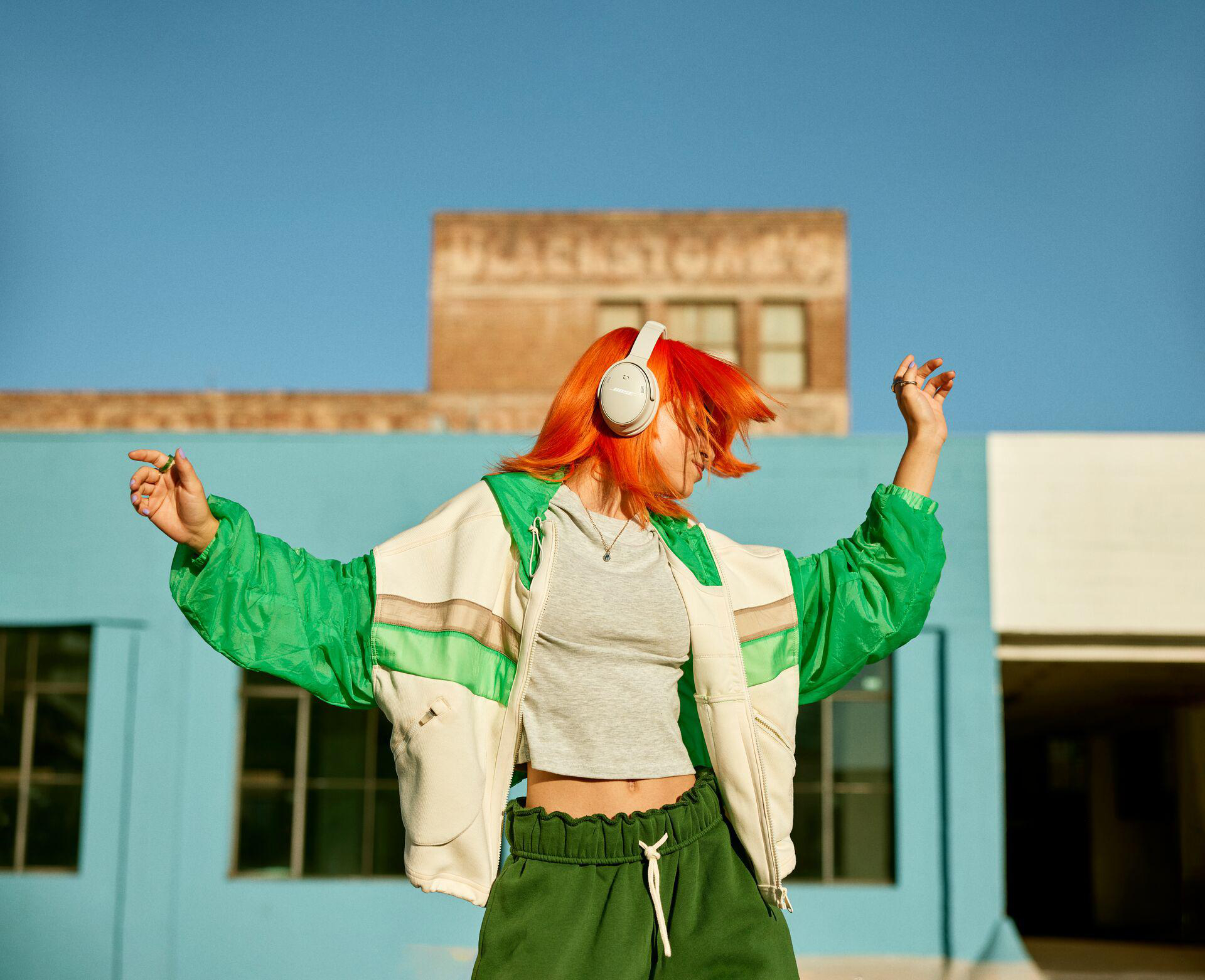 Frau mit orangefarbenen Haaren tanzt mit weißen Kopfhörern. Blauer Himmel im Hintergrund.