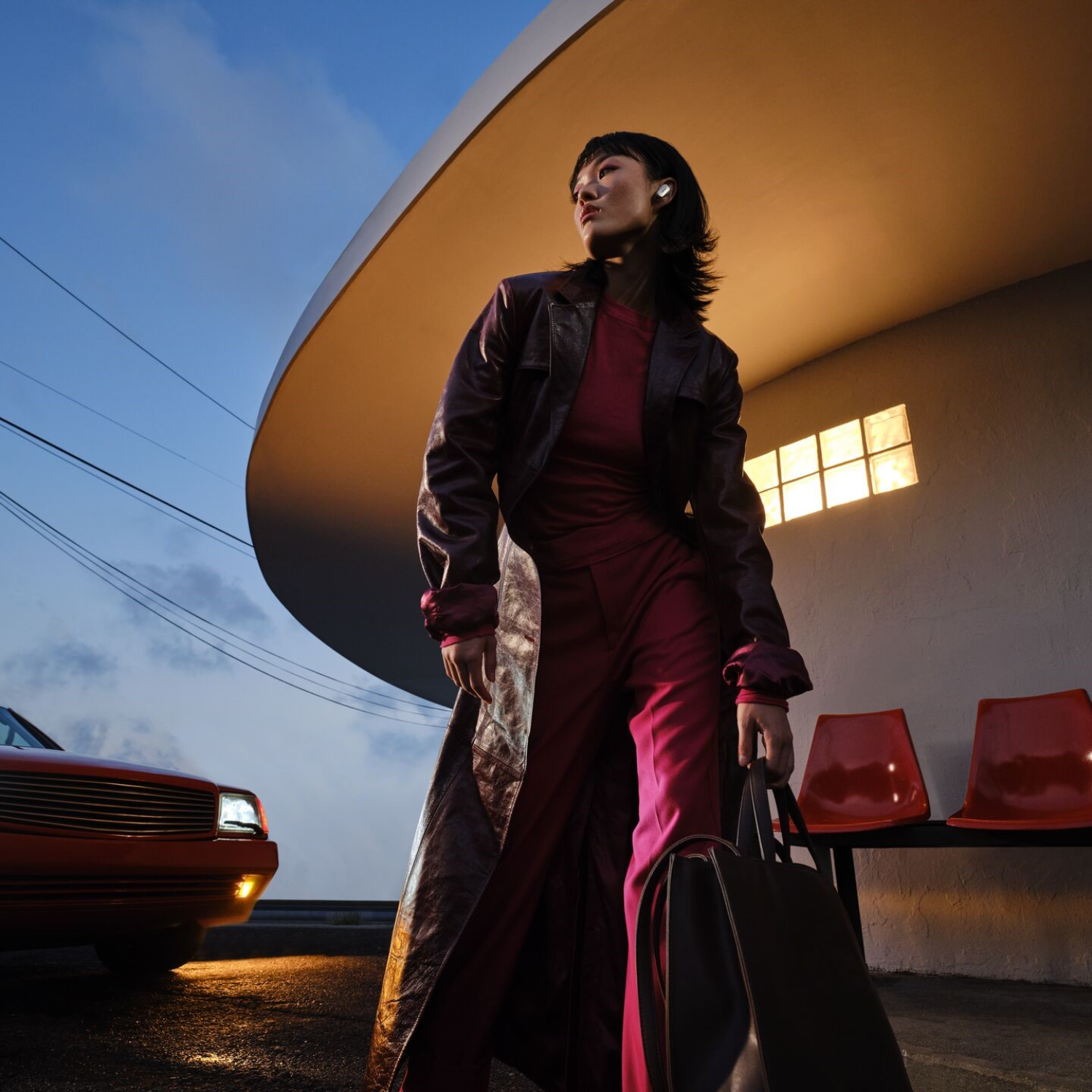Woman in red suit and leather coat at a bus stop with a red car nearby.