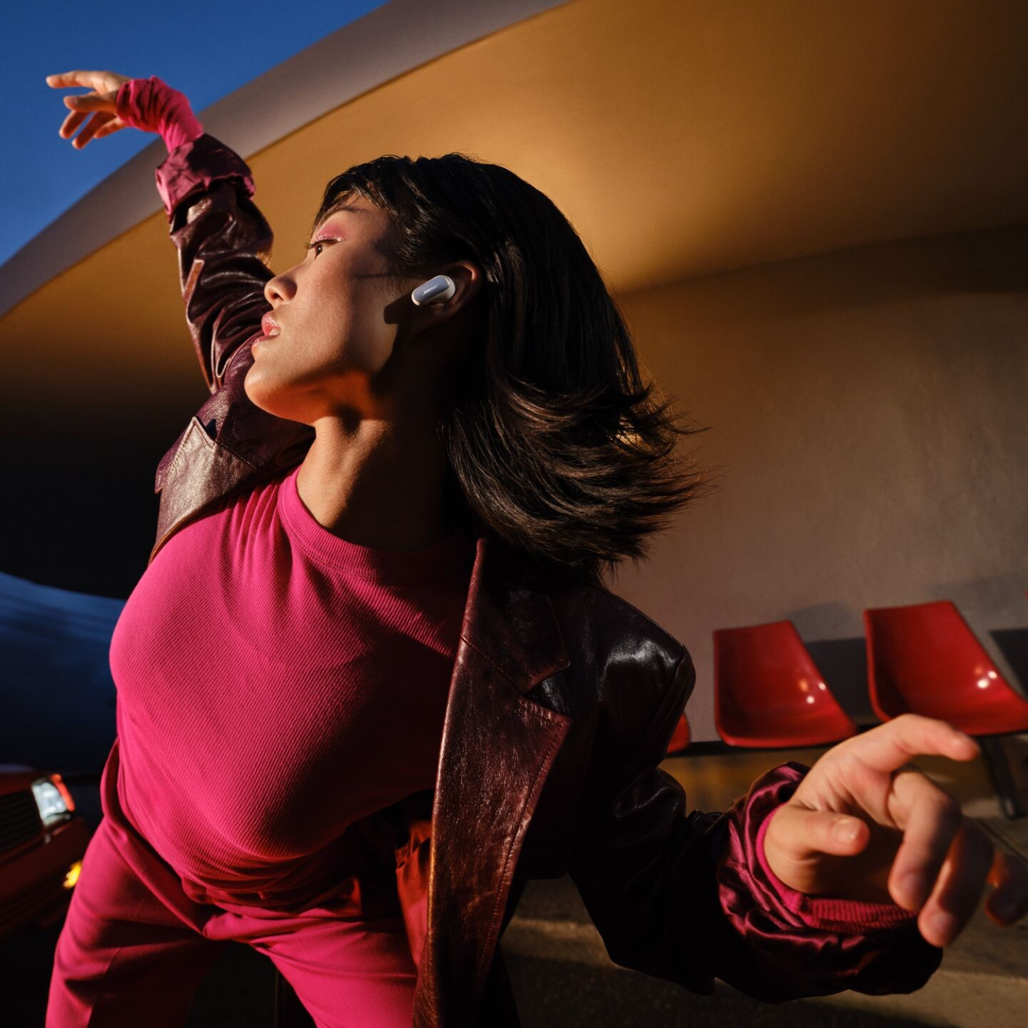 Woman dancing, wearing pink outfit, with earbuds. Sunset background, red chairs visible.