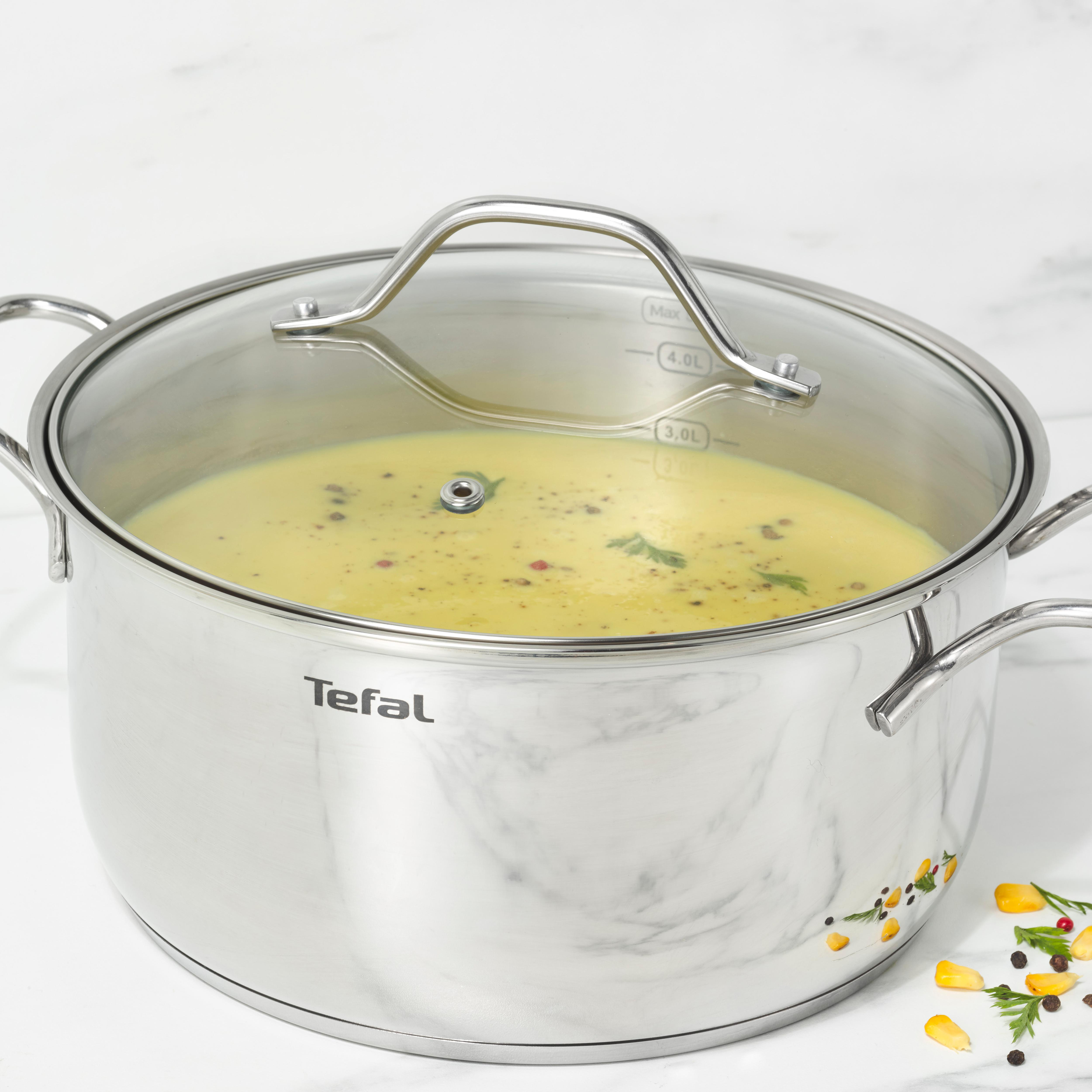 Ein Edelstahlkochtopf mit einem durchsichtigen Deckel, gefüllt mit gelber Suppe. Einige Zutaten verstreut.