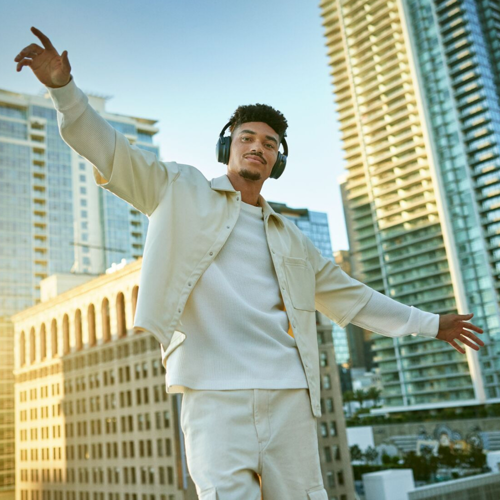 Young man with headphones, arms outstretched, standing in front of city buildings.