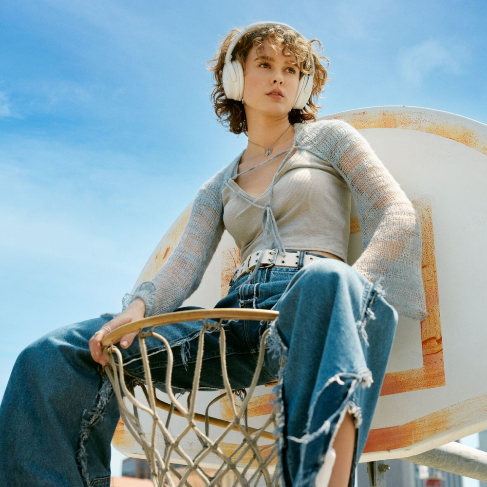 Woman with headphones sits on basketball backboard. Blue sky, denim jeans.