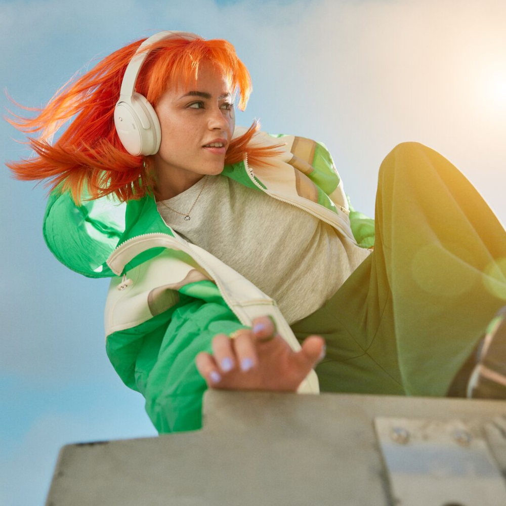 A woman with orange hair and white headphones is looking to the left, against a blue sky.
