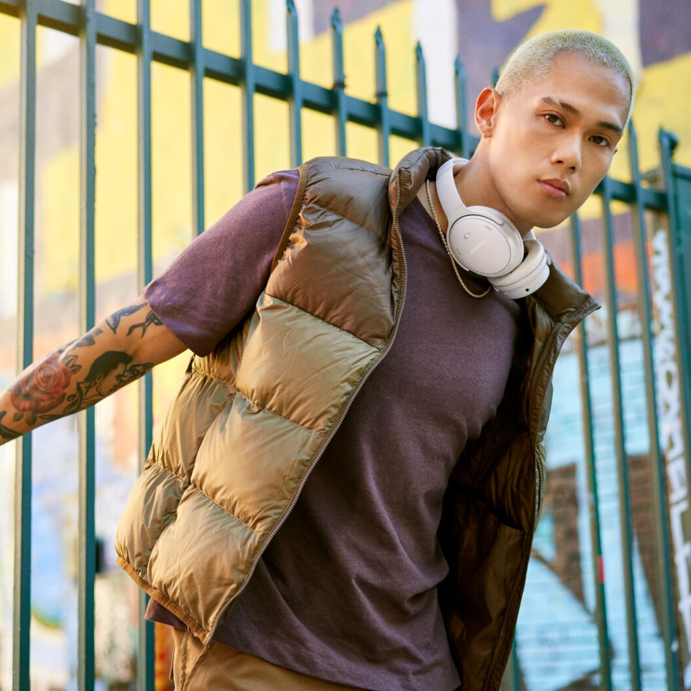A young man in a vest and headphones poses outdoors. There is a painted wall in the background.