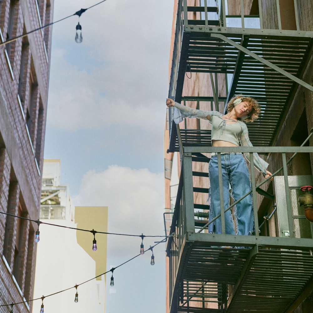 Woman on a fire escape, arms outstretched, buildings in the background, string lights.