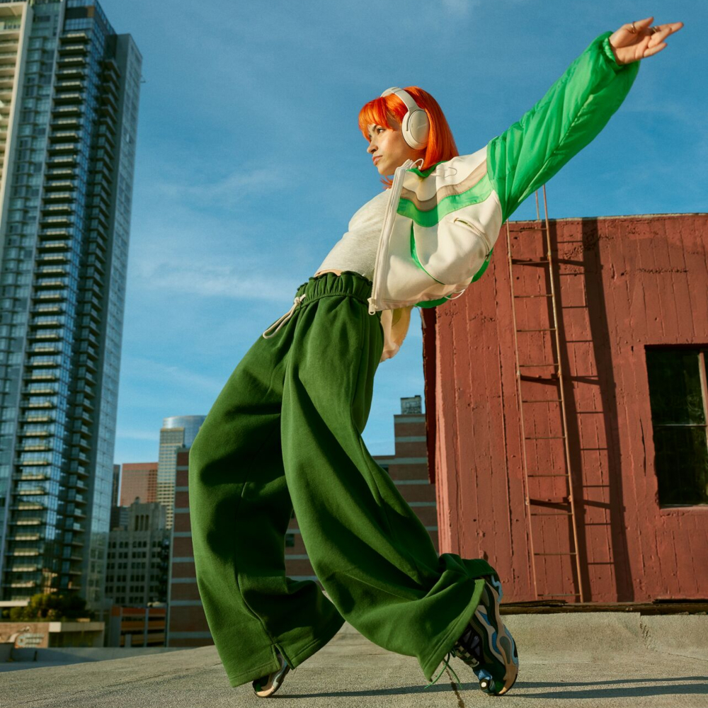 Woman with orange hair wearing headphones and green pants, on a rooftop, city view.