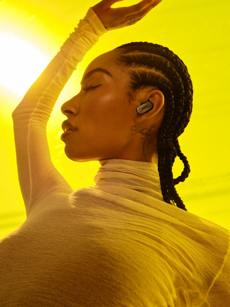 A woman with braids wearing a white top, wearing earbuds. Yellow background with bright sunlight.