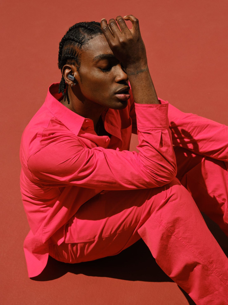 Man in red suit with earbuds, hand on face, red background.