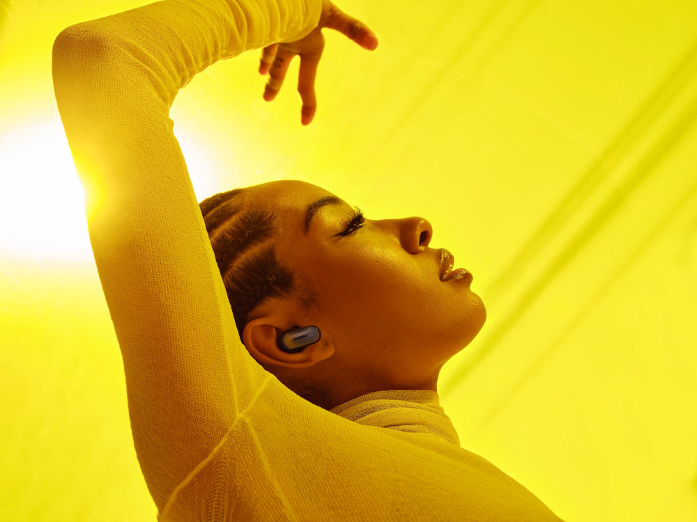 Woman with earbuds, arm raised, in front of a yellow backdrop.