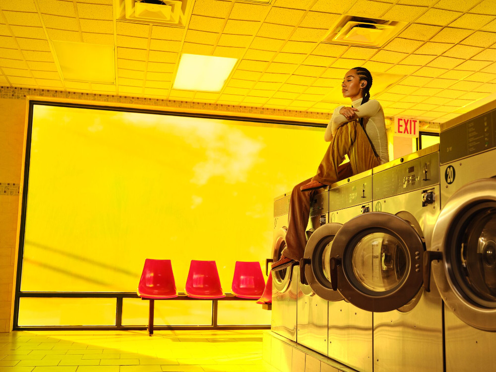A woman sits on washing machines. Bright yellow room, red chairs, and a yellow sky view.