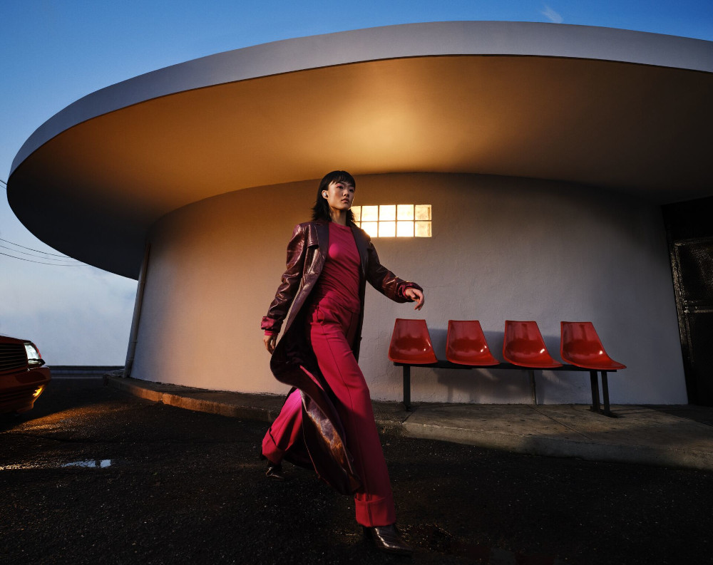Vrouw in roze outfit loopt langs een modern gebouw met rode stoelen.