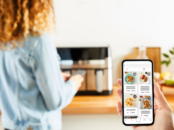 Persona sosteniendo un teléfono con una aplicación de recetas abierta frente a un electrodoméstico de cocina.