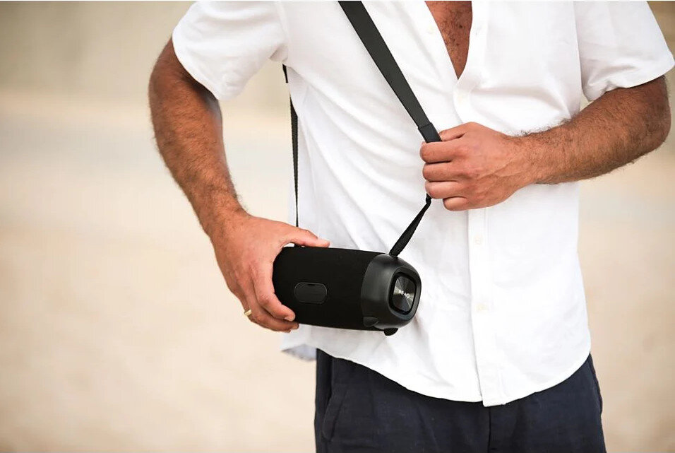 Mężczyzna w białej koszuli trzyma czarny głośnik z paskiem. Beżowe tło.