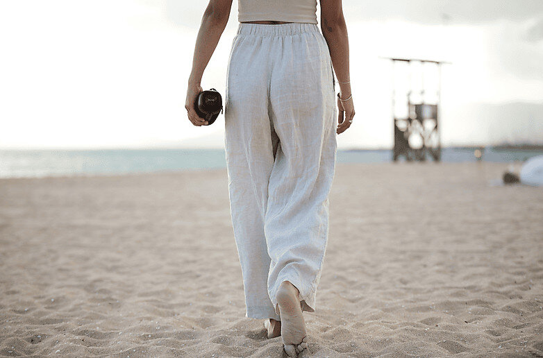 Kobieta w lnianych spodniach odchodzi na piaszczystej plaży, trzymając aparat, słoneczny dzień.