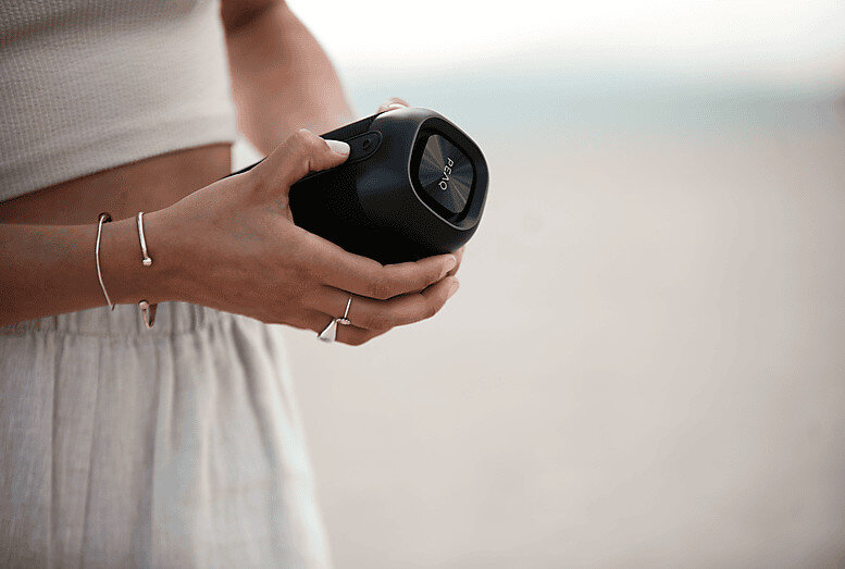 Osoba trzymająca czarny, cylindryczny głośnik z widoczną marką PEAQ na tle rozmytej plaży.