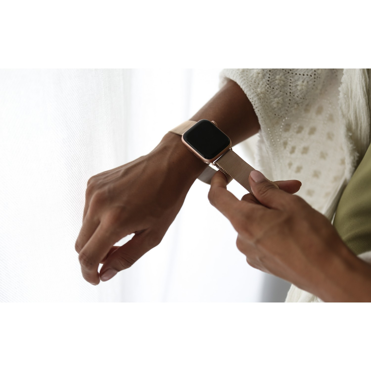 De hand van een vrouw houdt een gouden Apple Watch met een gouden mesh band vast, dicht bij een wit gordijn.