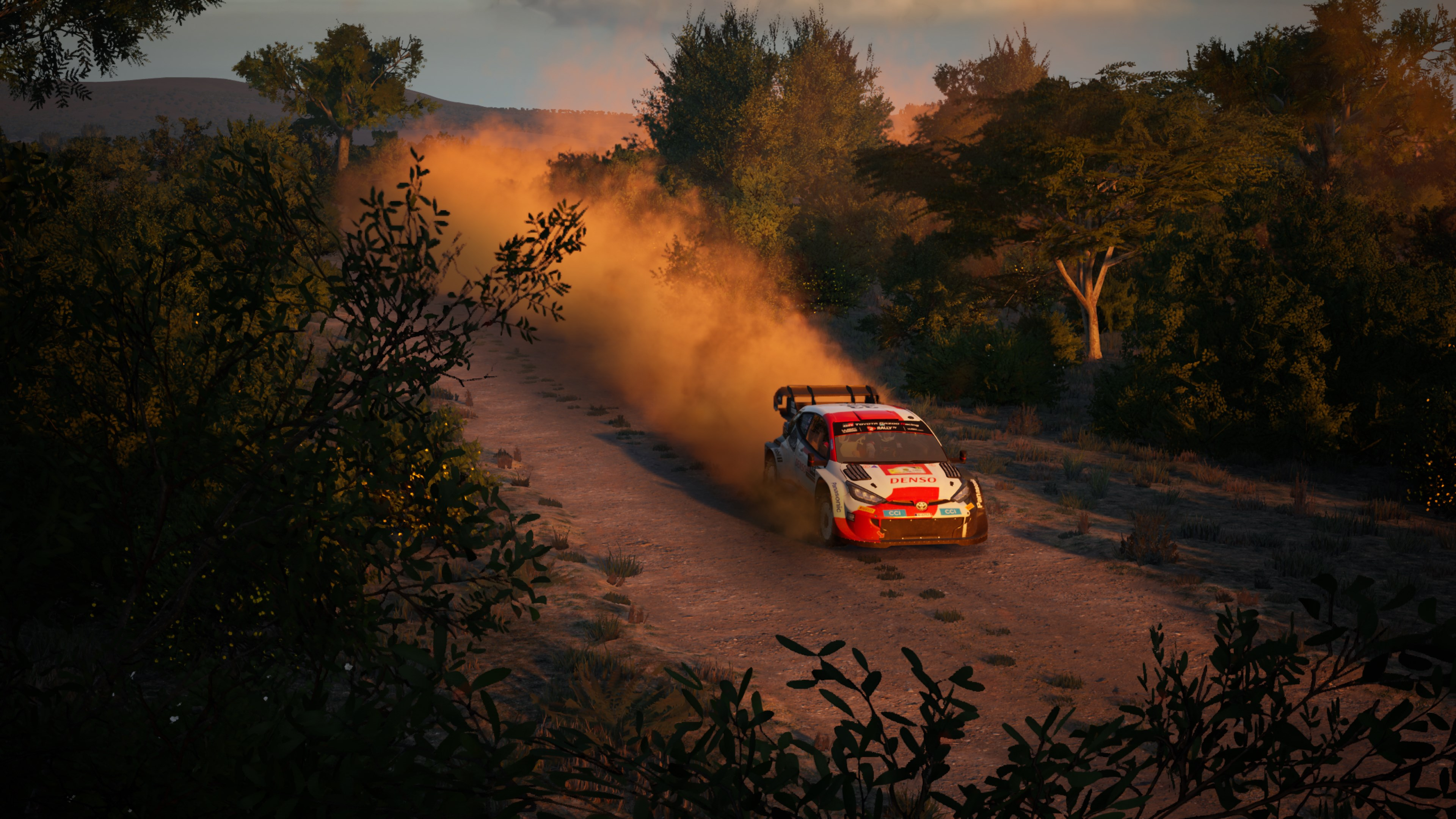 Une voiture de rallye soulève de la poussière sur un chemin de terre, entourée d'arbres, au coucher du soleil.