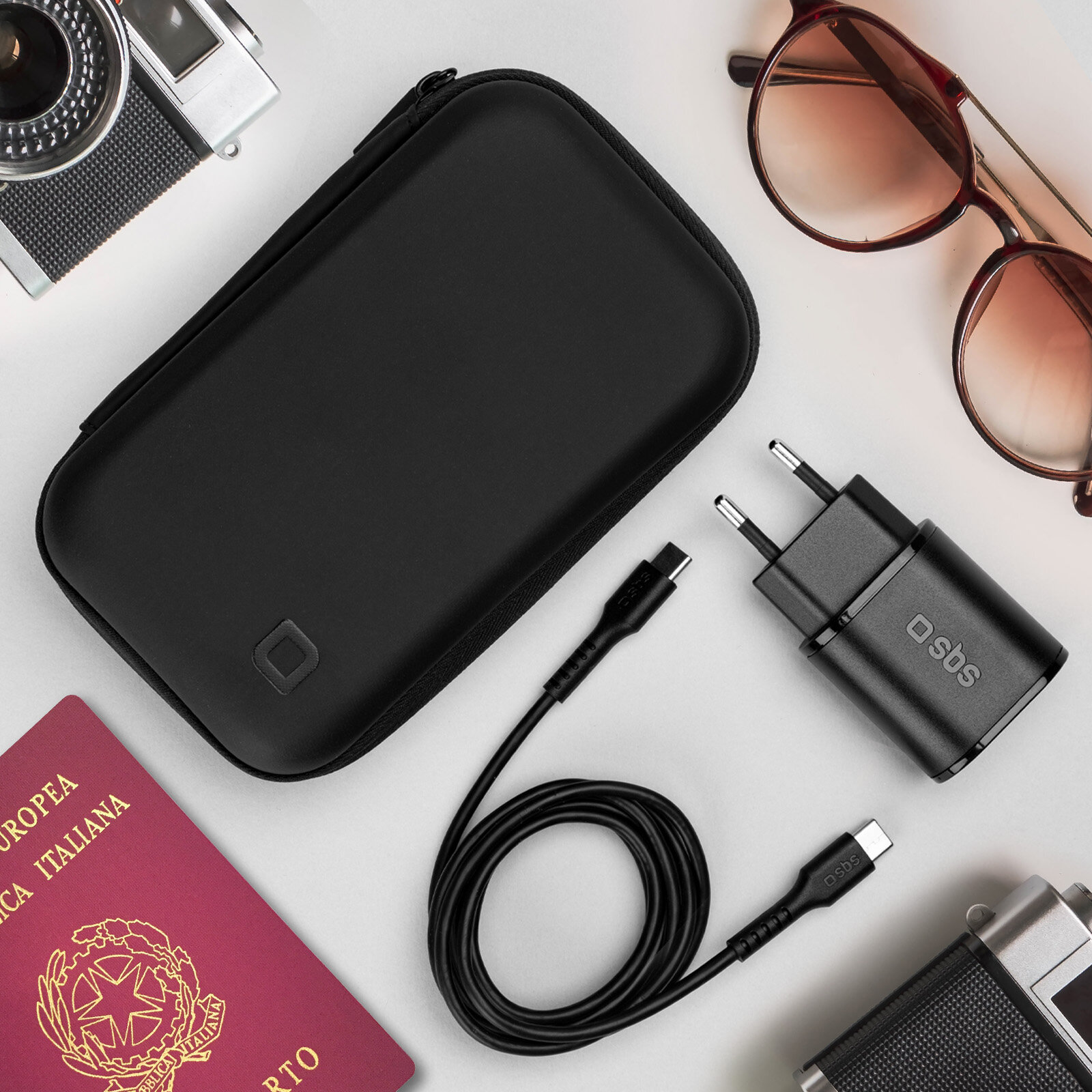 Czarny futerał podróżny, ładowarka, kabel, okulary przeciwsłoneczne i paszport na białej powierzchni.
