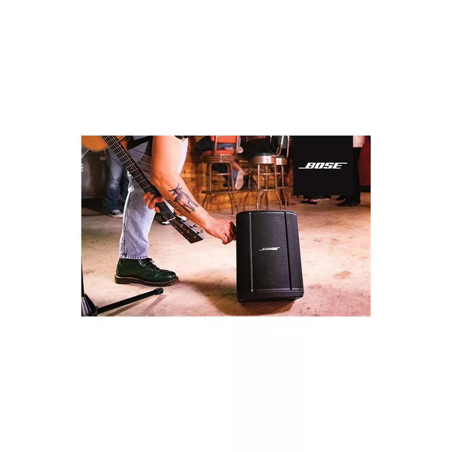 Man speelt gitaar, bij een zwarte speaker met het BOSE-logo. Een barscène op de achtergrond.