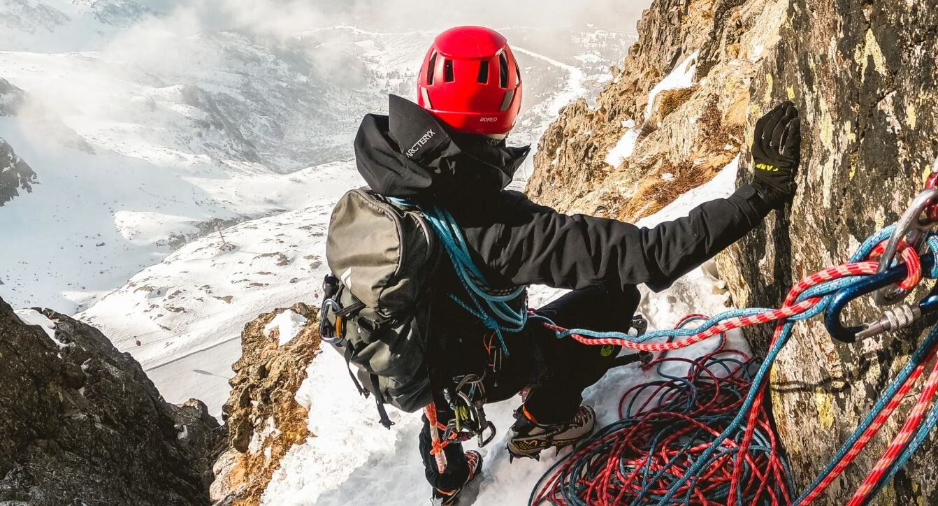 Osoba wspinająca się na zaśnieżoną górę, nosząca czerwony kask i sprzęt.