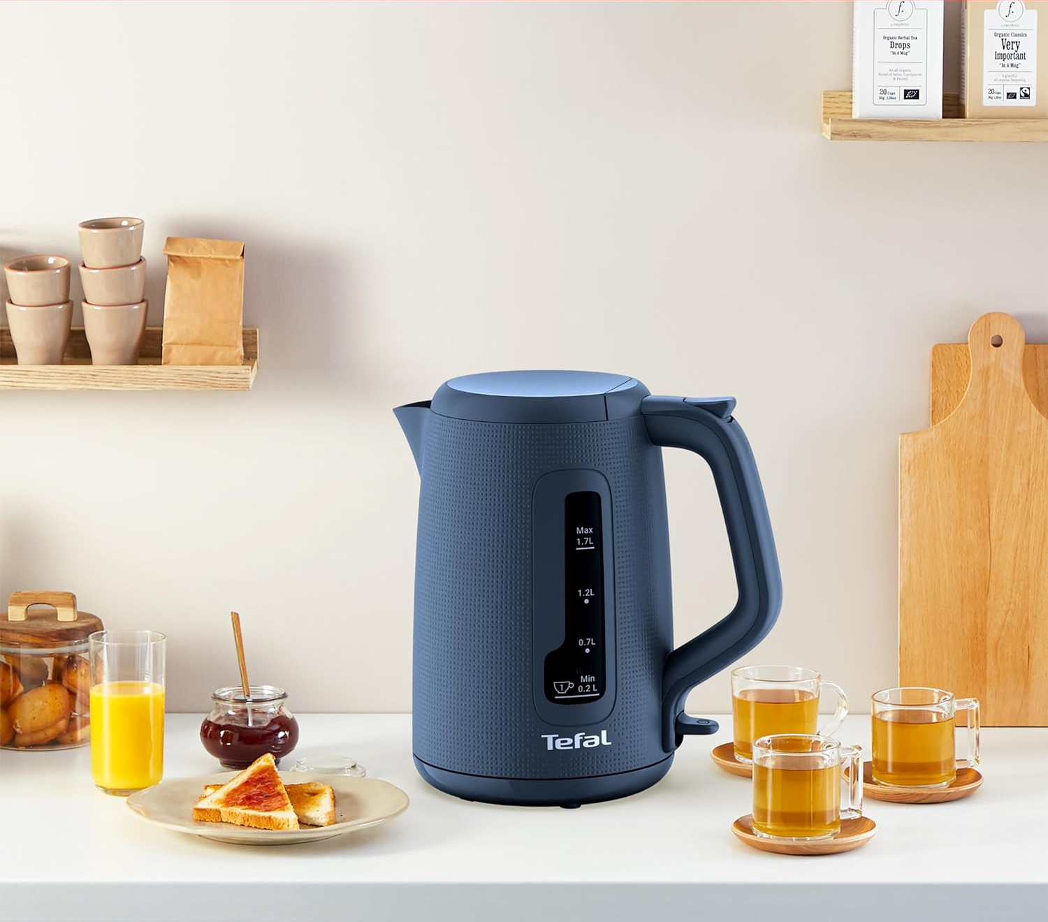Ein blauer Wasserkocher neben Toast mit Marmelade, Orangensaft und Teetassen auf weißer Theke.