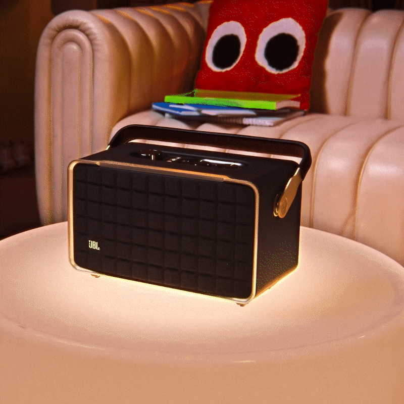 Schwarzer JBL-Lautsprecher auf einem Tisch. Rotes Kissen und Bücher im Hintergrund.