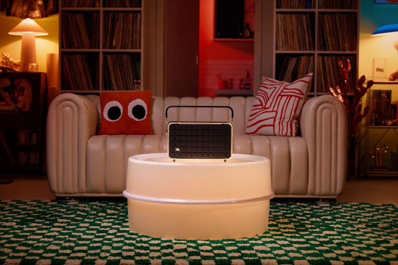 Ein Lautsprecher auf einem beleuchteten Tisch. Ein Sofa mit Kissen und Schallplatten im Hintergrund.