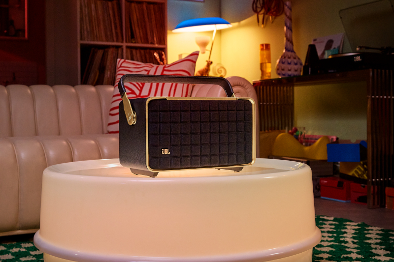 Ein schwarzer und goldener JBL-Lautsprecher steht auf einem weißen Tisch. Ein gemütliches Zimmer mit Sofa und Schallplatten im Hintergrund.