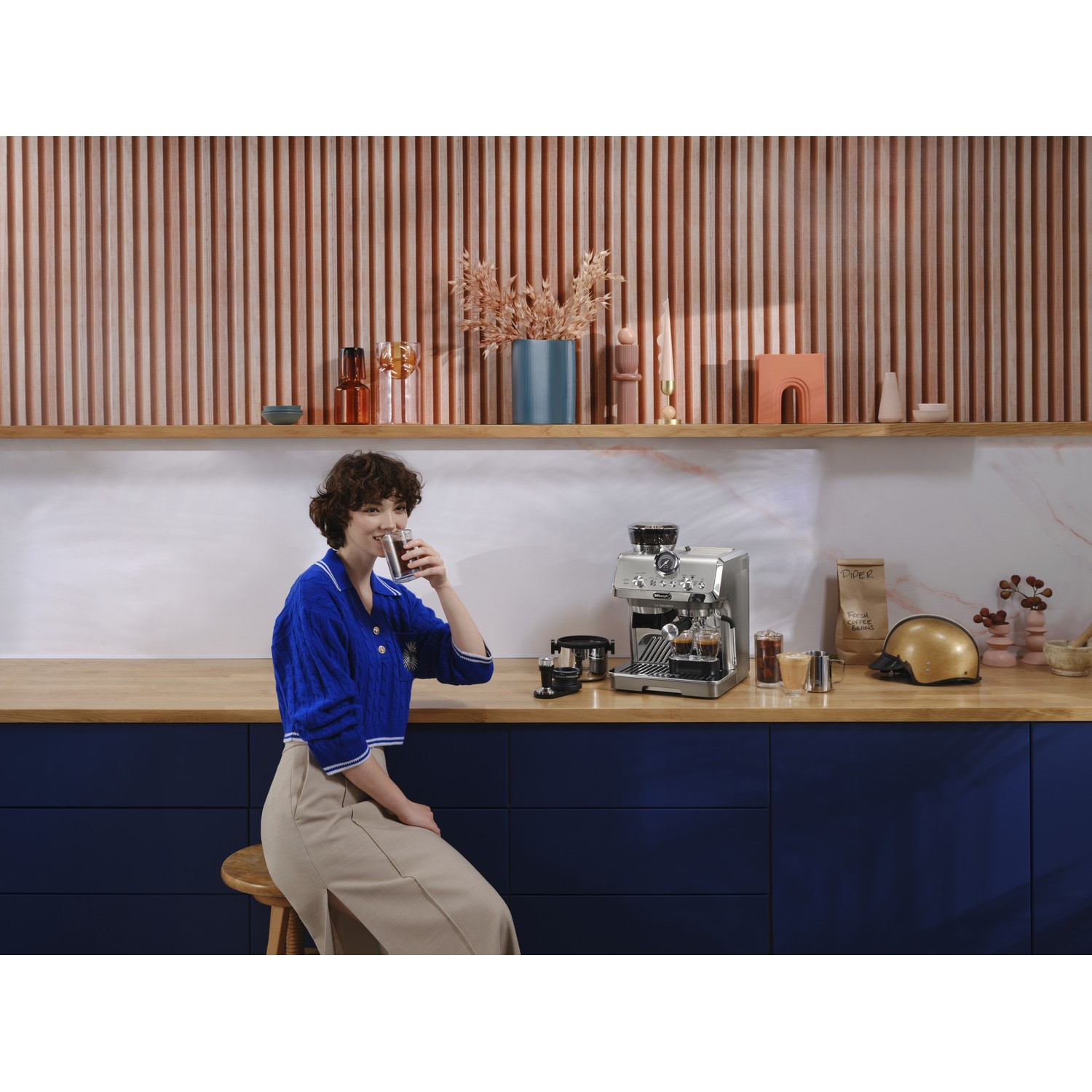 Vrouw drinkt uit een glas, zittend. Espressomachine en accessoires op het aanrecht. Houten en roze decor.
