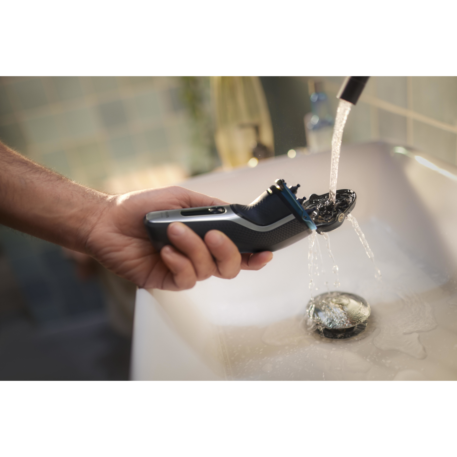 A hand holds a black and silver shaver above a sink with water flowing.