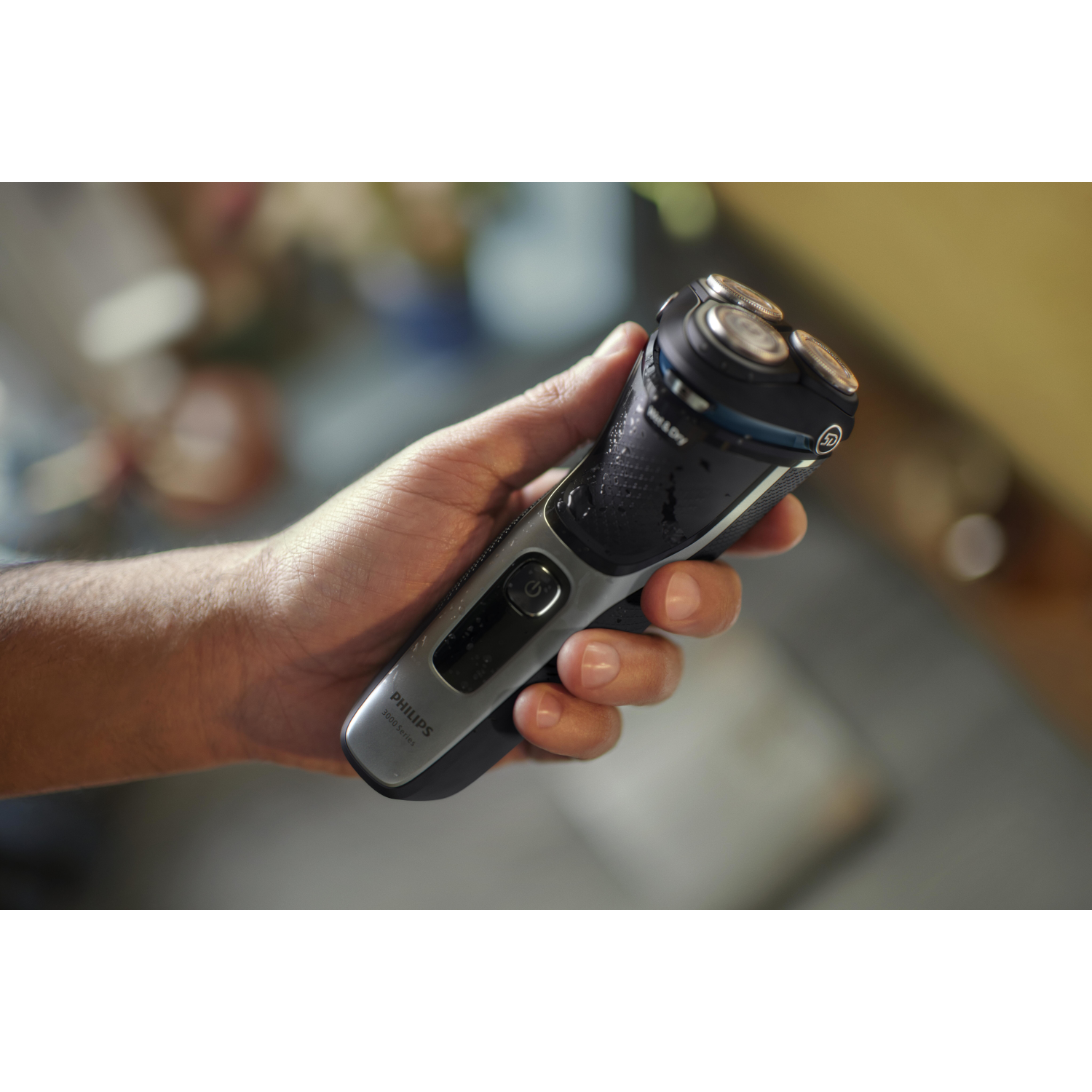 A hand holds a black and gray electric shaver, showcasing its three rotary heads and power button.