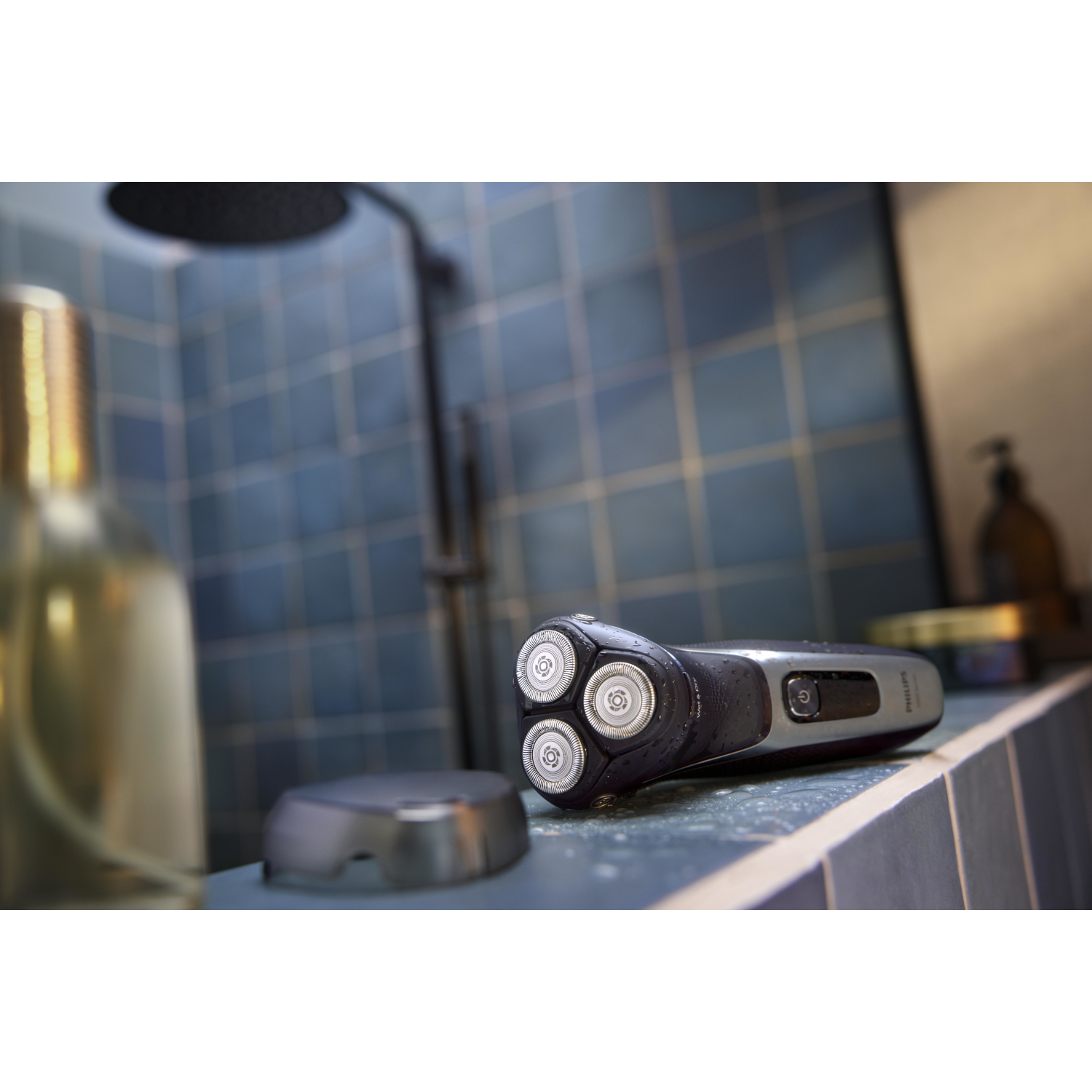 Electric shaver on a counter in a bathroom. Blue tiles and a showerhead are in the background.