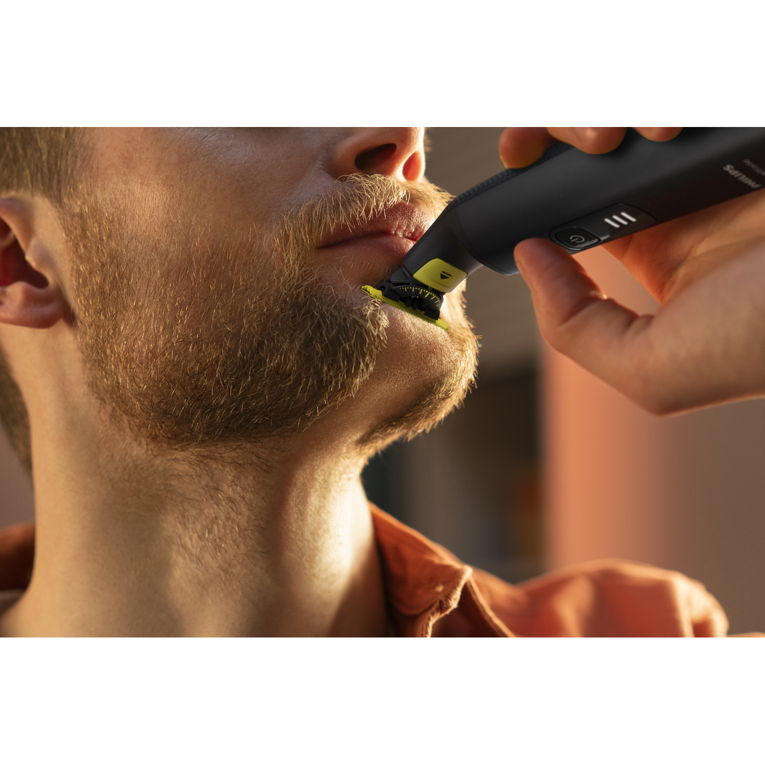 Man shaving his mustache with a trimmer, close up, with orange shirt.