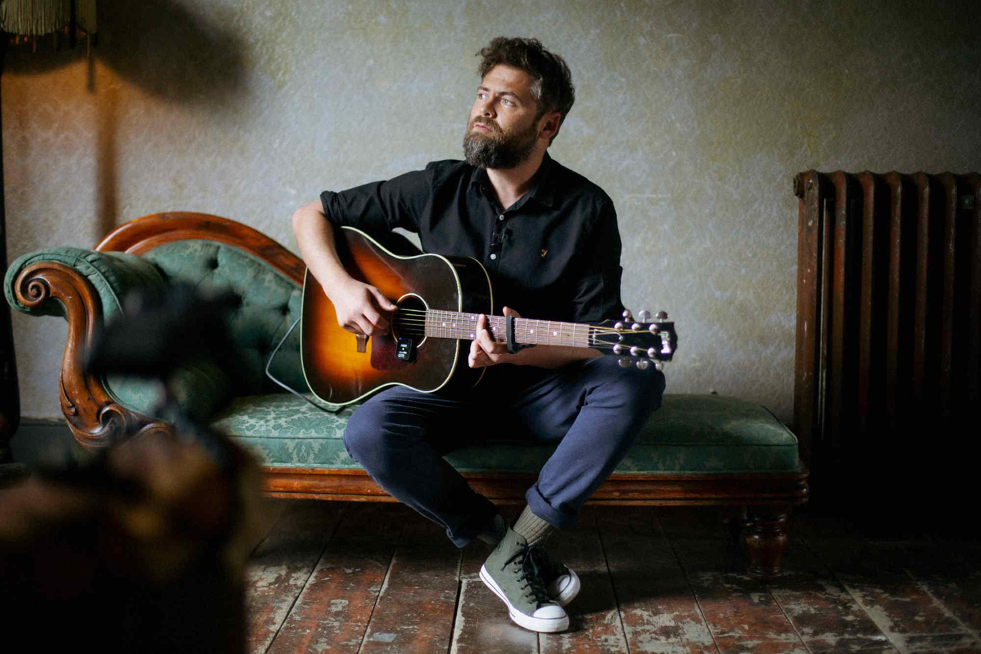 Ein Mann spielt Gitarre auf einem Sofa in einem dunklen Raum.