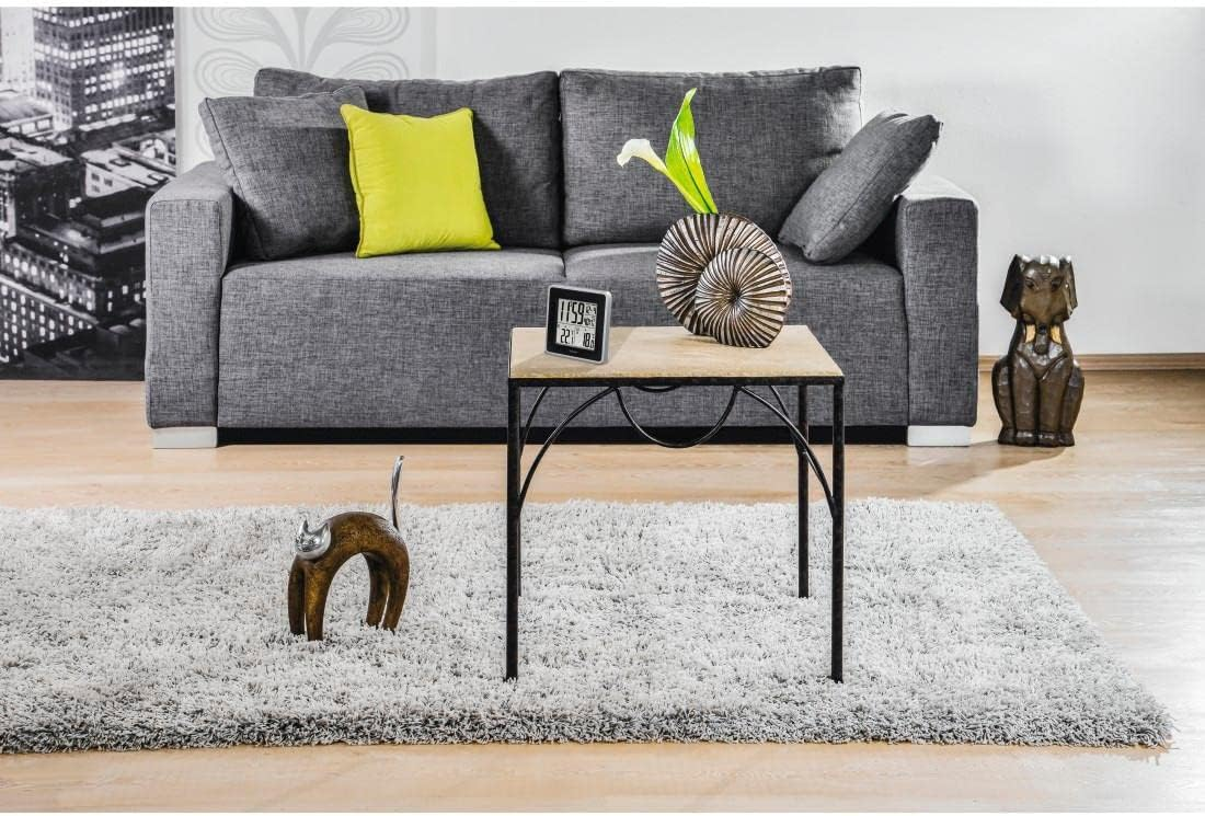 Sofá gris con cojines, mesa pequeña, alfombra y elementos decorativos. Escena urbana visible al fondo.