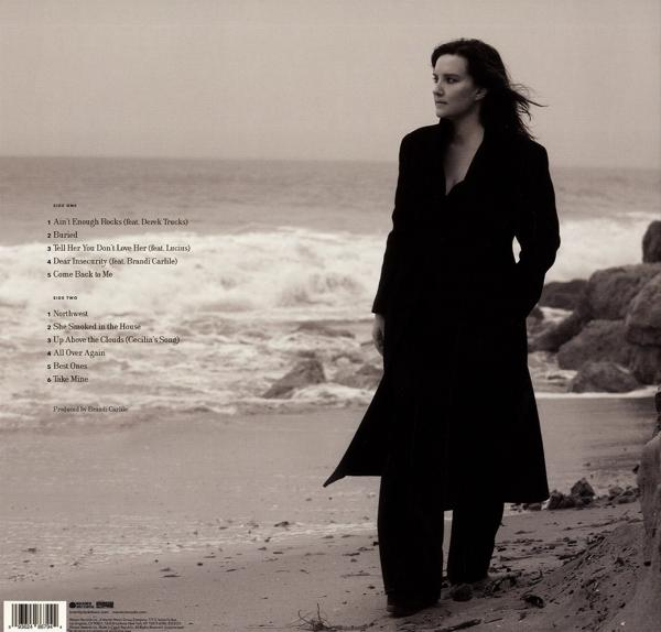 Frau in schwarzem Mantel steht am Strand, Blick nach rechts. Sand und Felsen im Hintergrund, bedeckter Himmel.