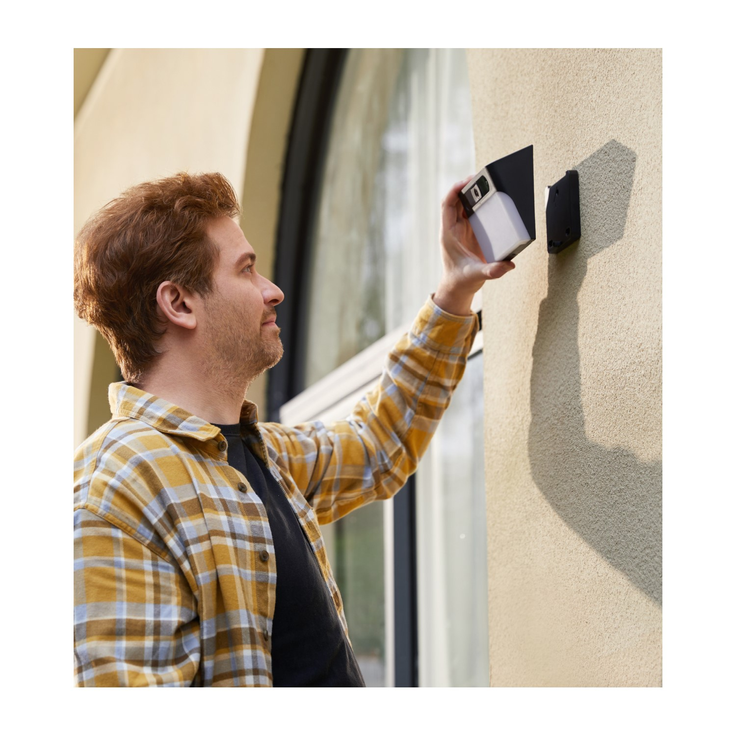 Een man installeert een wit en zwart apparaat op een beige muur naast een raam.