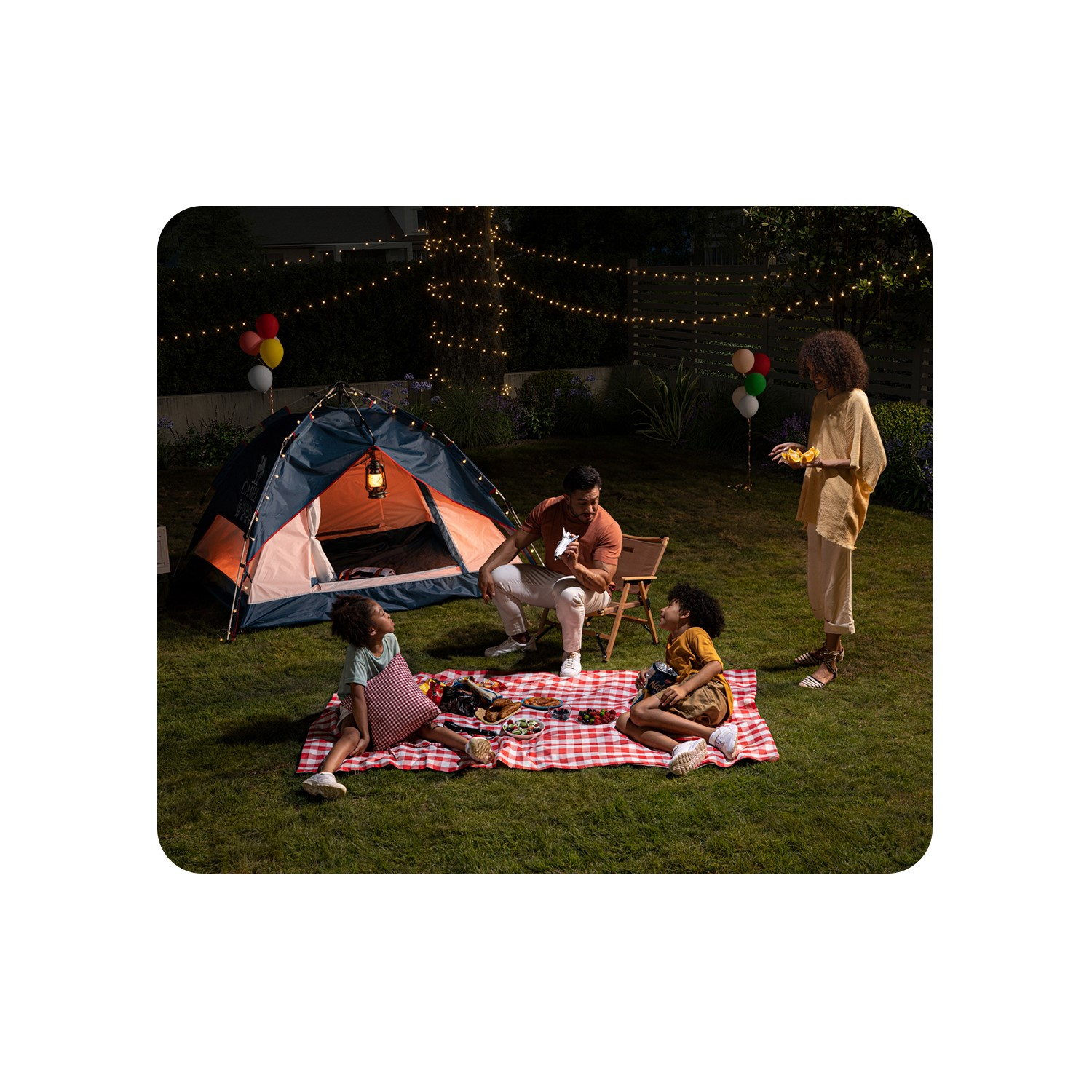 Pique-nique familial dans un jardin. Tente, couverture de pique-nique, ballons et guirlandes lumineuses. Personnes assises, mangeant et parlant.